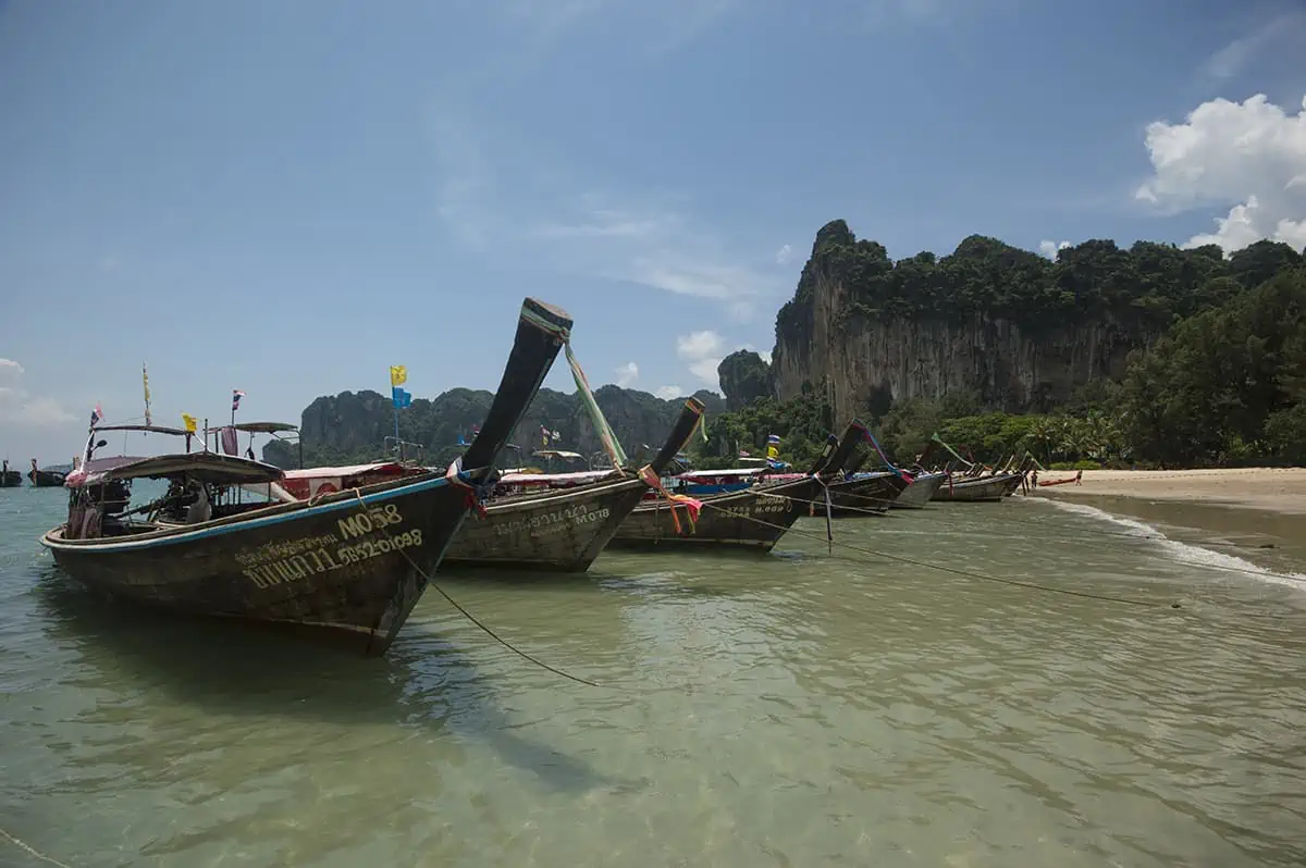 Ao Nang To Railay Beach: The ULTIMATE Guide 2025 10 Longtail boats anchored just offshore in the shallow waters of Railay Beach with cliffs towering behind.