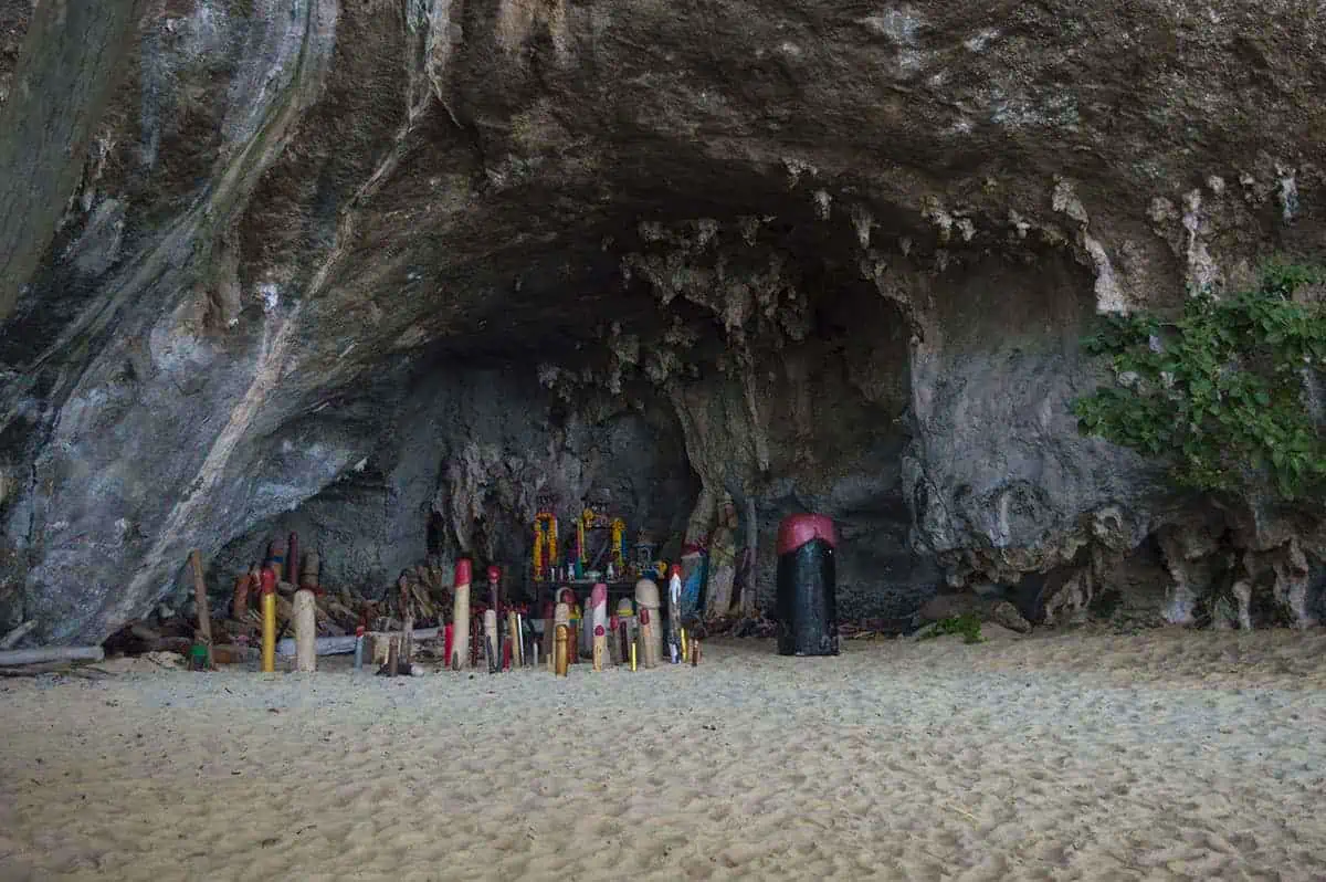 Ao Nang To Railay Beach: The ULTIMATE Guide 2025 14 Phra Nang Cave shrine on Railay Beach, featuring numerous colorful wooden phallic offerings on the sandy floor beneath a rocky overhang.