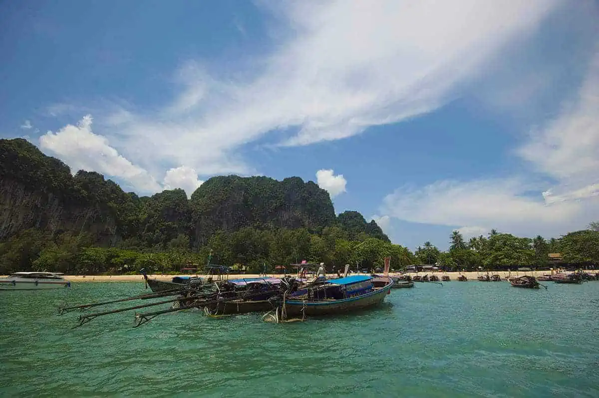 Ao Nang To Railay Beach: The ULTIMATE Guide 2025 21 Longtail boats floating near the shore of Railay Beach with lush green cliffs and clear water under a blue sky.