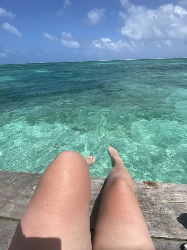 Bacalar To Caye Caulker: The ULTIMATE Travel Guide 2025 15 View of a Tasha's legs dangling off a wooden dock, with her feet hovering above the clear waters of Caye Caulker.