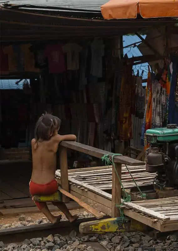 Battambang Bamboo Train: Is It Still Worth Visiting? 2025 14 A young child sits at the edge of a bamboo train cart, parked in front of a market stall with hanging clothes and in the background.
