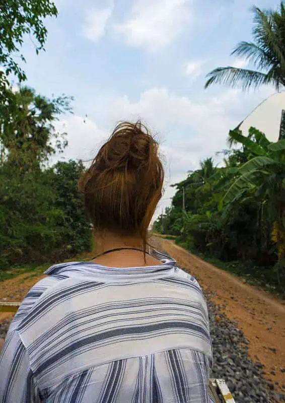 Battambang Bamboo Train: Is It Still Worth Visiting? 2025 18 tasha amy with her back to the camera sits on a battambang bamboo train riding along the railway, surrounded by palm trees and jungle.