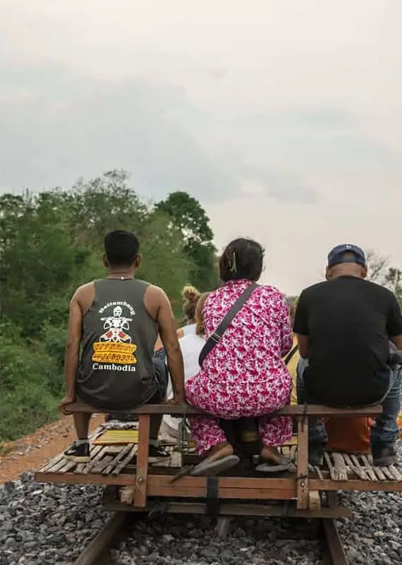 Battambang Bamboo Train: Is It Still Worth Visiting? 2025 15 View from behind several locals riding together on a single bamboo train cart, including a man in a tank top with “Cambodia” written on the back.