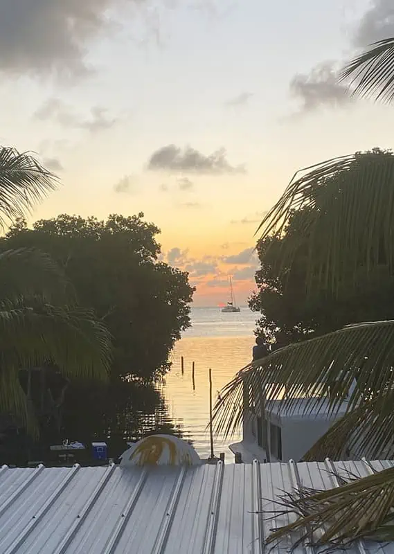 A serene sunset view from a rooftop in Belize, showcasing hues of orange and yellow blending into the sky. Palm fronds frame the top of the image, and the calm water reflects the sunset, with a sailboat moored in the distance. This peaceful scene captures the tranquil beauty of Belize in April.