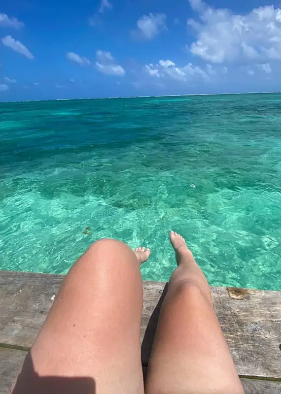 Offering a first-person perspective, this image shows the bare legs of a person sitting on a weathered wooden dock, looking out over the shallow, crystal-clear waters of Belize in April. The water is so transparent that the sandy bottom is visibly detailed.