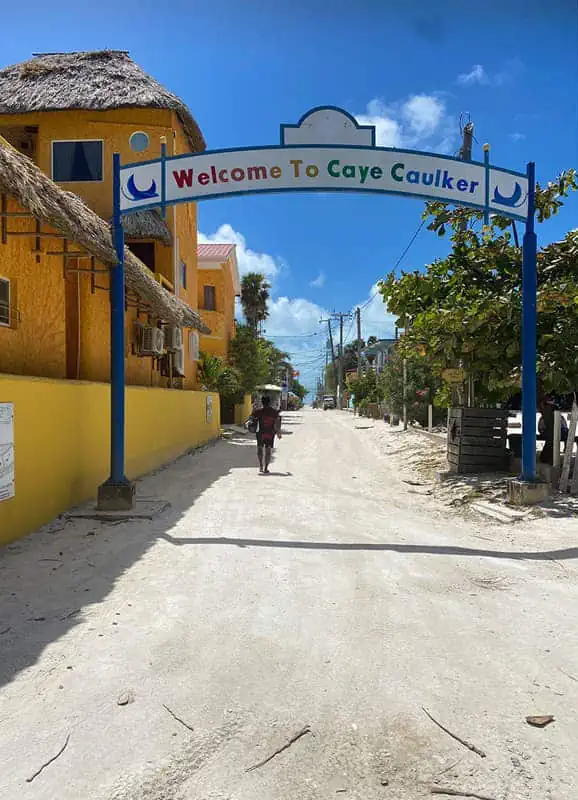 The main sandy street on Caye Caulker, Belize, under a bright blue sky. An arched sign reading "Welcome to Caye Caulker" spans the road, framed by tropical yellow and blue buildings on either side, welcoming visitors to the island.
