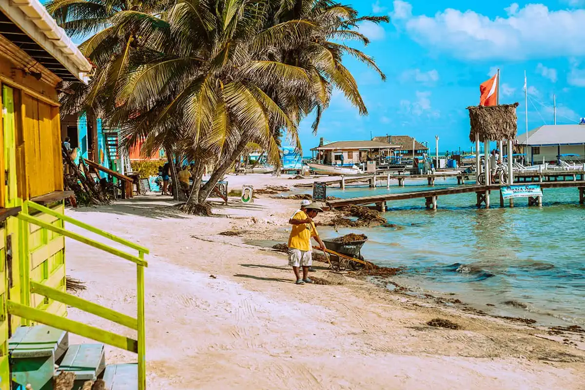 A local Belizean in a yellow shirt walking along a beachside path with wooden docks and colorful buildings, typical of a June day in Belize.