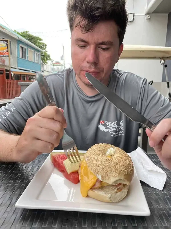 A man enjoying a savory breakfast bagel with melon slices, depicting a modern twist to a Belizean breakfast in a casual outdoor setting.