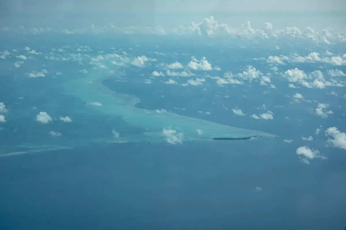 The image captures a wider aerial perspective of the same atoll in Belize, featuring scattered clouds above and the clear turquoise waters that surround the lush green atoll.