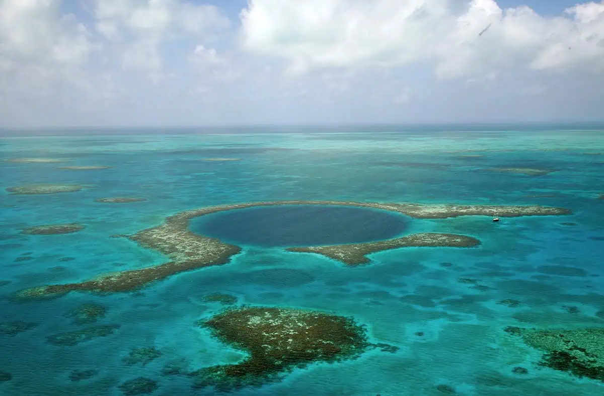 Atolls In Belize: The 3 Best Paradise Spots Worth Visiting 1 Aerial view of the famous Great Blue Hole at lighthouse atoll in Belize surrounded by shallow coral reefs and a solitary boat near its edge, illustrating the scale and beauty of this natural wonder.