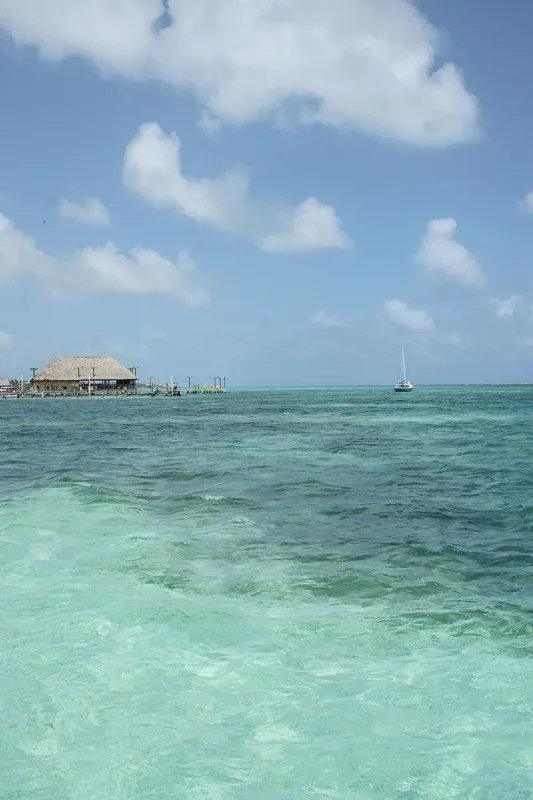 Best Beaches In Ambergris Caye: For Swimming, Snorkeling, Tanning & More 19 A view of a calm sea with a thatched hut on a pier and a sailboat in the distance under a vast blue sky, highlighting the serene beauty of Ambergris Caye's beaches.