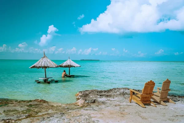 Two thatched umbrellas in the shallow turquoise waters of Ambergris Caye, with a person relaxing underneath and wooden lounge chairs on the beach, representing one of the best beaches in Ambergris Caye.