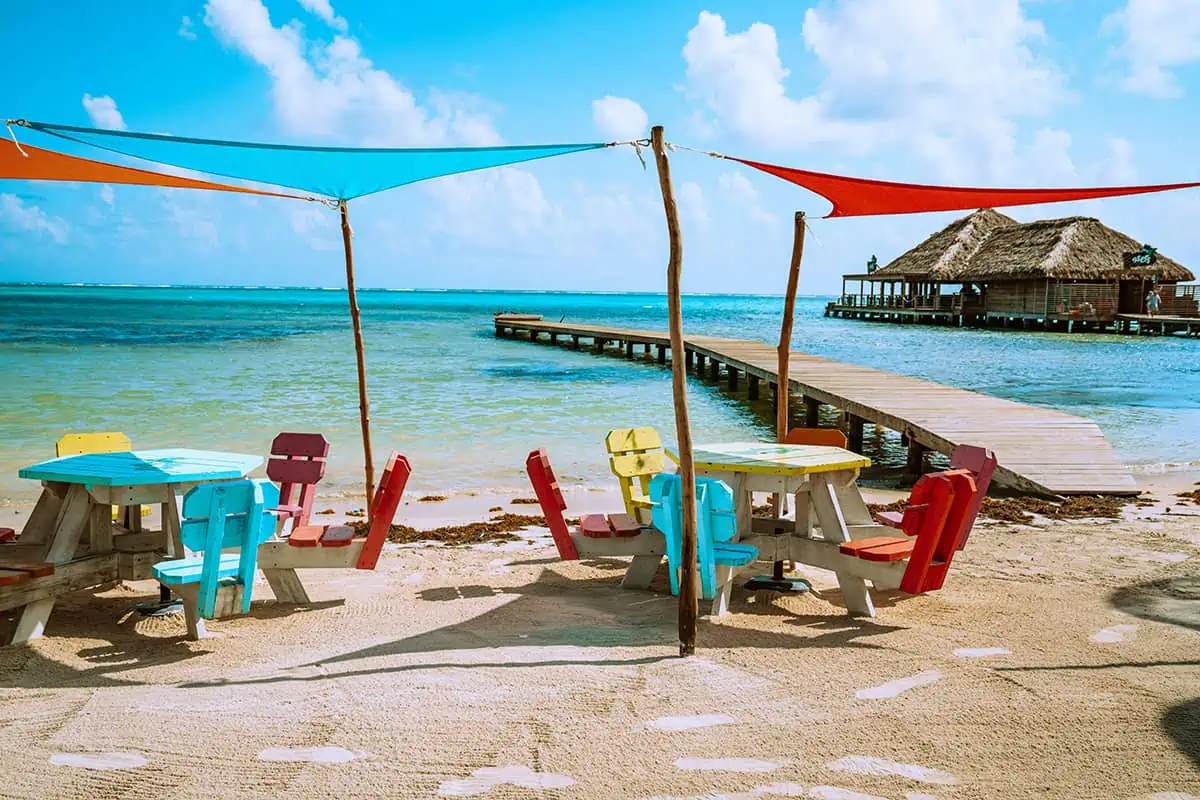 Best Beaches In Ambergris Caye: For Swimming, Snorkeling, Tanning & More 6 Colorful beach chairs and a picnic table under shade sails on the sandy shores of Ambergris Caye, with a view of the calm turquoise waters and a thatched pier in the background, inviting relaxation at one of the best beaches.