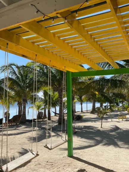 Best Beaches In Ambergris Caye: For Swimming, Snorkeling, Tanning & More 10 A view under a bright yellow pergola with swings, looking out to a sandy beach with palm trees and clear skies, a relaxing corner on Ambergris Caye.