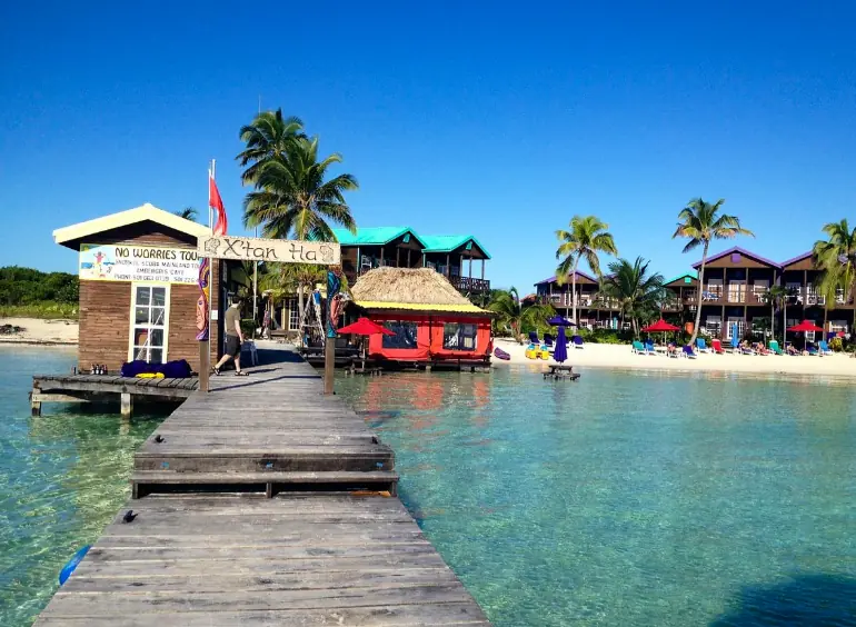 Best Beaches In Ambergris Caye: For Swimming, Snorkeling, Tanning & More 11 A wooden pier leading to vibrant beachfront businesses and palm trees, with the calm sea inviting visitors to explore the best beaches in Ambergris Caye.