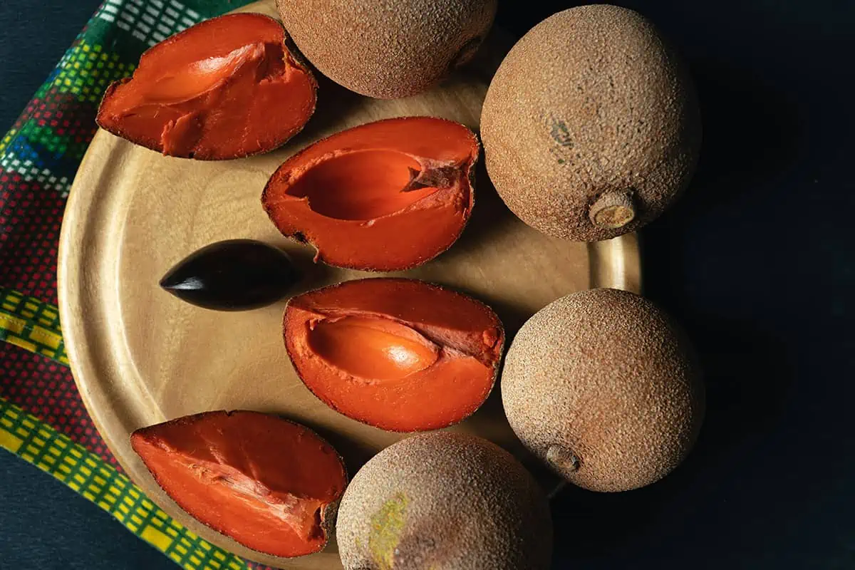 Fruits In Belize: The BEST 24 Fruits Worth Trying On Your Visit 19 A wooden plate displaying open mamey sapote fruits with reddish-orange flesh and a glossy black seed, a tropical fruit often enjoyed in Belize.
