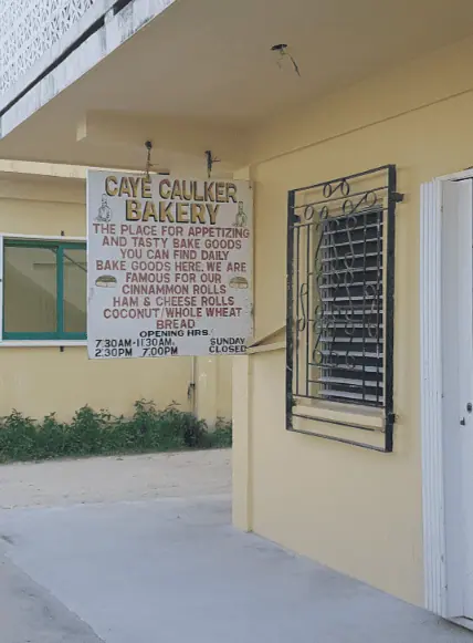 BEST Restaurants On Caye Caulker Worth Trying 2025 12 Exterior view of Caye Caulker Bakery, prominently featured as one of the best restaurants on Caye Caulker. The bakery's facade is painted a warm yellow, and a large white sign hangs above a barred window, detailing the delicious offerings such as daily fresh cinnamon rolls and whole wheat bread. This bakery is highlighted as the place for 'appetizing and tasty bake goods' in Caye Caulker, open daily with posted hours for convenient visitor access.
