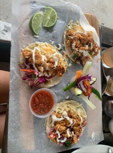 BEST Restaurants On Caye Caulker Worth Trying 2025 15 Close-up of three shrimp tacos served on a paper-lined tray at Barrier Reef Sports Bar & Grill, one of the best restaurants on Caye Caulker. Each taco is filled with crispy fried shrimp, fresh cabbage slaw, and drizzled with creamy sauce, accompanied by a lime wedge and a small bowl of salsa, providing a colorful and appetizing presentation