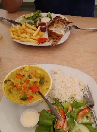 BEST Restaurants On Caye Caulker Worth Trying 2025 19 A table at Chef Juan's Kitchen and Pastries, renowned as one of the best restaurants on Caye Caulker, showcasing two plates of hearty meals. The first plate features grilled fish with a side of fries and a garden salad, and the second plate offers a curry dish with mixed vegetables served alongside white rice and a green salad, highlighting the fresh, local ingredients used in their cuisine.