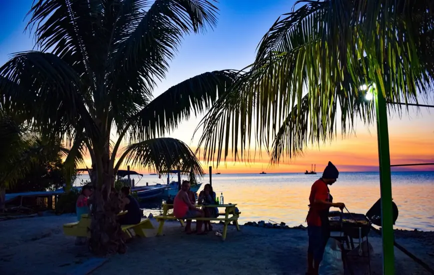 BEST Restaurants On Caye Caulker Worth Trying 2025 21 An enchanting evening scene at Dee N D's Waterside Eatery, widely regarded as one of the best restaurants on Caye Caulker. The photograph captures diners seated at bright yellow picnic tables under the swaying palm trees along the beachfront. A cook in a red shirt is barbecuing by the shore as the sun sets in the background, painting the sky in vivid hues of orange and blue, reflecting off the calm Caribbean waters. This setting exemplifies a perfect tropical dining experience on Caye Caulker.