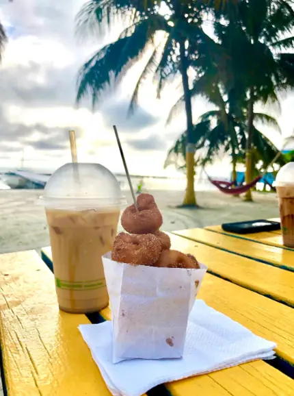 BEST Restaurants On Caye Caulker Worth Trying 2025 8 A delightful morning snack scene at Ice N Beans, one of the best restaurants on Caye Caulker for a quick bite. A paper cone filled with freshly made donuts, dusted with cinnamon sugar, is paired with a tall plastic cup of iced coffee on a yellow picnic table. The table is set against a backdrop of a sandy beach and palm trees, offering a quintessential tropical setting.