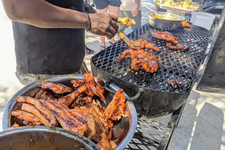 BEST Restaurants On Caye Caulker Worth Trying 2025 14 A bustling street food scene at Chef Kareem's UnBelizeable Lunch, known as one of the best restaurants on Caye Caulker for authentic local cuisine. The photo shows a chef, deftly handling various pieces of juicy, barbecued chicken and ribs over a grill, with a bowl of marinade at hand for basting. The grill is filled with deliciously charred meats, enticing passersby with its smoky aroma. This image captures the vibrant street food culture that is a hallmark of Caye Caulker’s culinary offerings.