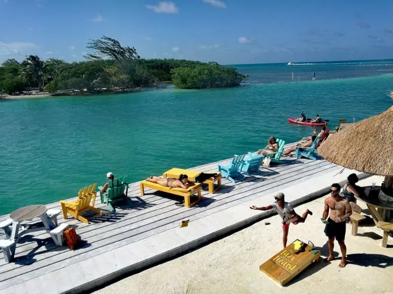 BEST Restaurants On Caye Caulker Worth Trying 2025 17 A vibrant, sunny day at Lazy Lizard, one of the best restaurants on Caye Caulker, captured from a bustling wooden pier. The image showcases a group of people enjoying the clear turquoise waters and serene views. Several lounge chairs and colorful wooden benches provide ample seating for guests to relax and soak in the sun. A few individuals are engaging in casual activities, adding to the lively atmosphere of this popular waterside destination.