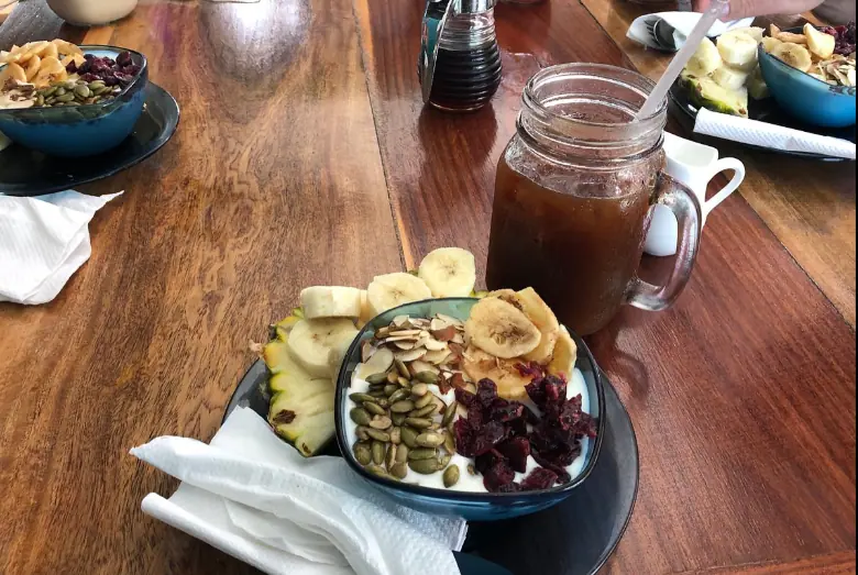 BEST Restaurants On Caye Caulker Worth Trying 2025 10 A healthy and vibrant breakfast spread at Namaste Cafe, renowned as one of the best restaurants on Caye Caulker for healthy dining. The image displays a bowl of yogurt topped with sliced bananas, pineapple, pumpkin seeds, and dried cranberries. Adjacent to it is a mason jar filled with iced tea, reflecting a refreshing start to a day. The meal is set on a polished wooden table, adding warmth to the wholesome and inviting atmosphere of the cafe