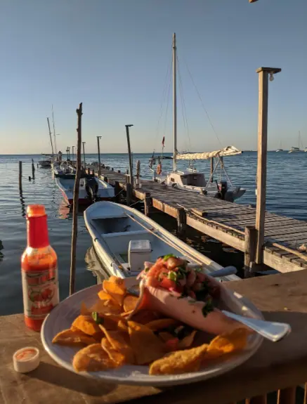 BEST Restaurants On Caye Caulker Worth Trying 2025 25 A picturesque dining experience at Pelican Sunset Bar, one of the best restaurants on Caye Caulker, featuring a plate of shrimp cocktail and fried plantains. The dish is served on a rustic wooden table overlooking a pier with moored boats and the calm Caribbean Sea, capturing the essence of a perfect seaside meal at sunset