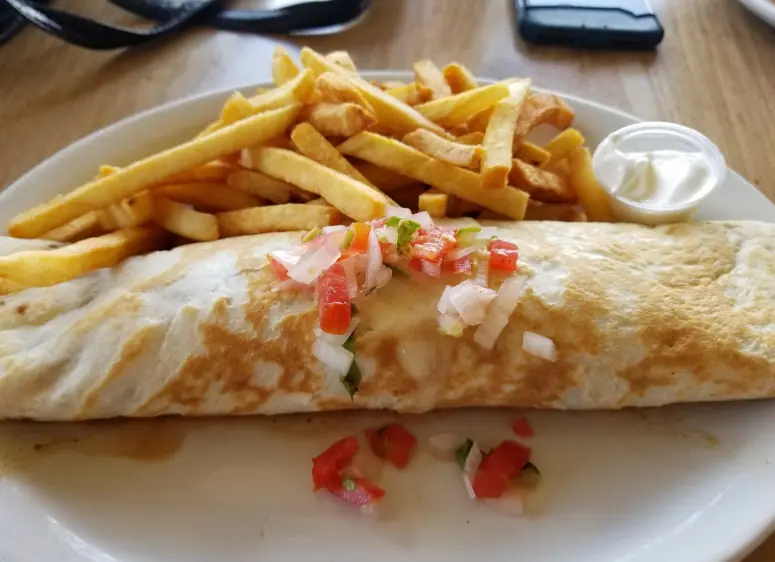 BEST Restaurants On Caye Caulker Worth Trying 2025 18 A classic lunch serving at Rainbow Grill & Bar, popularly known as one of the best restaurants on Caye Caulker. The plate features a large, golden-brown wrap topped with fresh pico de gallo, served alongside a generous portion of crispy French fries and a side of sour cream. This dish is presented on a simple white plate, symbolizing the straightforward yet delicious offerings of the restaurant, ideal for a satisfying meal after exploring the island
