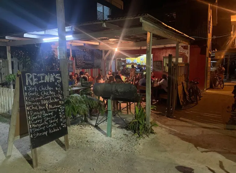 BEST Restaurants On Caye Caulker Worth Trying 2025 22 A lively evening scene at Reina's, a popular outdoor dining spot on Caye Caulker. The image captures a rustic, open-air restaurant bustling with diners seated at simple wooden tables. A chalkboard menu stands at the entrance, listing an array of dishes such as Jerk Chicken and Coconut Curry Chicken, illuminated by overhead string lights that add a cozy glow to the setting. A large, barrel-style grill is active in the background, with a chef preparing meals, enhancing the casual, communal atmosphere of the restaurant
