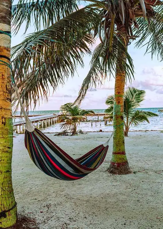 The 31 BEST Things To Do In Caye Caulker: Ultimate List! 36 A vibrant, multicolored hammock hanging between two palm trees on a sandy beach in Caye Caulker. The backdrop features a calm sea with a wooden pier stretching out into the water, evoking a sense of peace and tropical tranquility.