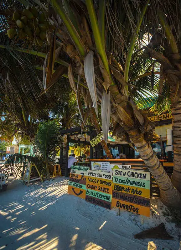 The 31 BEST Things To Do In Caye Caulker: Ultimate List! 32 A cozy beachside eatery in Caye Caulker, with hand-painted signs listing menu items like stew chicken, tacos, pupusas, and seafood dishes. The restaurant is shaded by large palm trees, offering a laid-back atmosphere perfect for enjoying local cuisine.