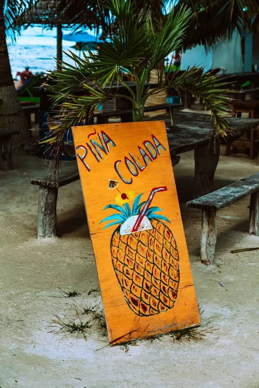 The 31 BEST Things To Do In Caye Caulker: Ultimate List! 31 A colorful wooden sign painted with a large pineapple and the words "Piña Colada," placed on sandy ground near rustic picnic tables and palm trees. This sign, near a beach bar, invites visitors to enjoy a refreshing tropical drink.