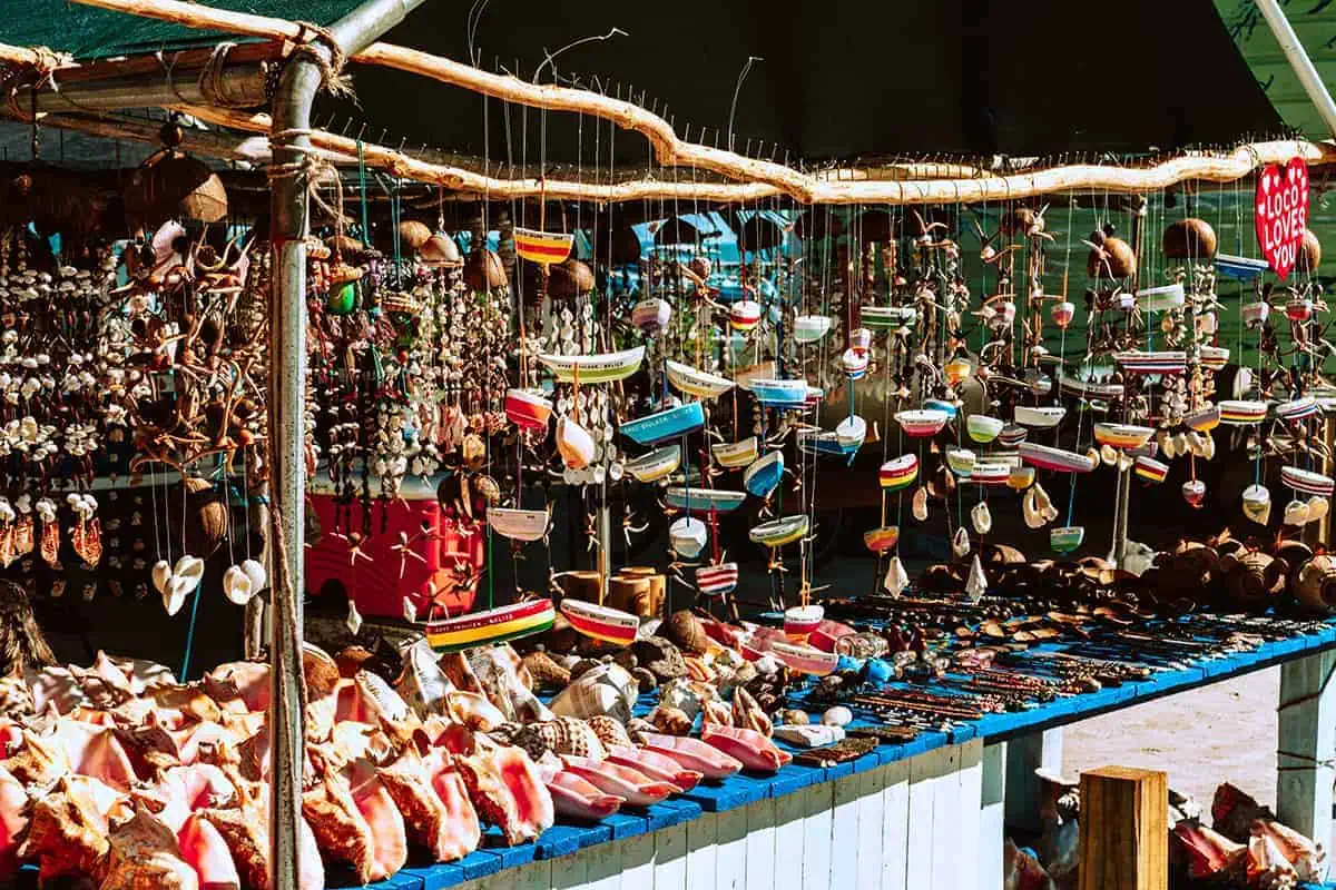 The 31 BEST Things To Do In Caye Caulker: Ultimate List! 22 A bustling market stall in Caye Caulker, filled with an assortment of handcrafted items such as shell ornaments, miniature wooden boats, and colorful jewelry. The stall is shaded by a green canopy, providing a vibrant and inviting shopping experience for tourists.