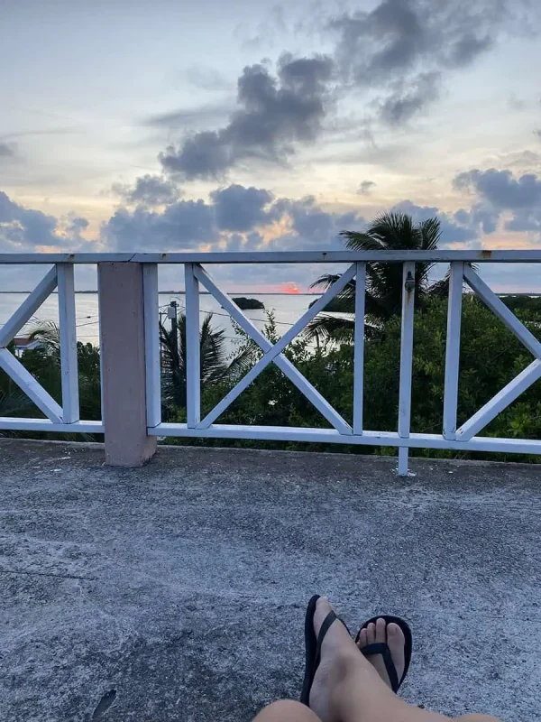 41 Best Things To Do In Ambergris Caye For The Ultimate Holiday 34 A tranquil scene at dusk on Ambergris Caye with a person's legs resting on a balcony railing, showcasing their relaxed posture and flip-flops with a stunning view of the ocean and a vibrant sunset in the background.