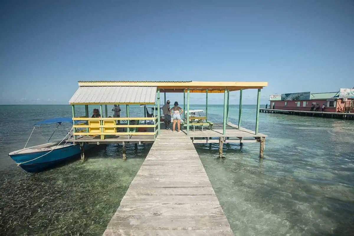 41 Best Things To Do In Ambergris Caye For The Ultimate Holiday 8 A rustic wooden dock in Caye Caulker extending into the turquoise sea with a boat tied up and people enjoying the view, a common relaxing activity on the island.