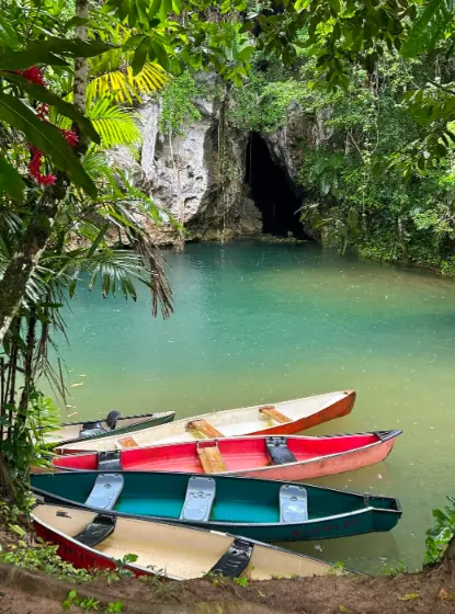 25+ Best Things To Do In San Ignacio, Belize (2025) 27 Colorful canoes docked by the edge of a serene, turquoise river. The river is surrounded by lush tropical vegetation, and the barton creek cave entrance is visible in the background, offering a picturesque and peaceful setting for outdoor activities in San Ignacio.
