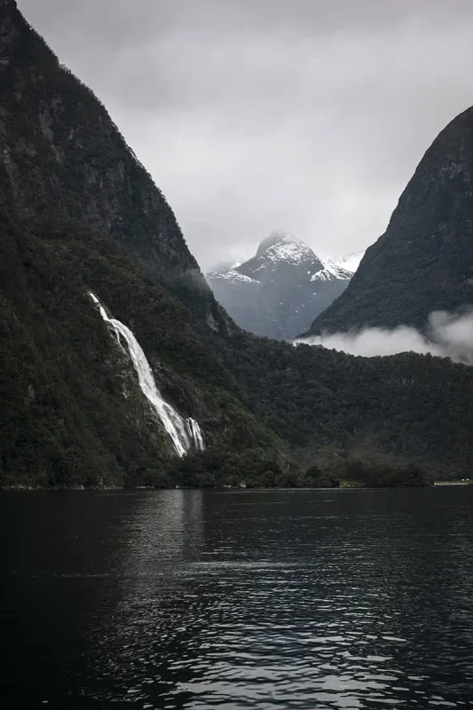 Bowen Falls In Milford Sound: The ULTIMATE Guide 2025 6 A close-up view of Lakes Bowen Falls in Milford Sound, with the waterfall dramatically descending between two steep, forested cliffs. Snow-capped peaks loom in the background, partially obscured by mist and low clouds. The calm, reflective water below captures the stark contrast of the bright waterfall against the dark, verdant landscape.