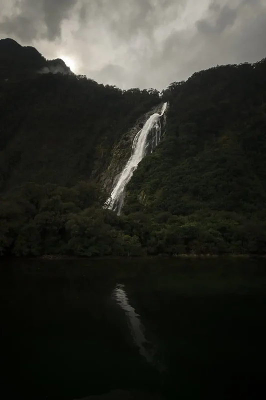Bowen Falls In Milford Sound: The ULTIMATE Guide 2025 5 The magnificent Bowen Falls is framed by dense green foliage and rugged mountain terrain. The waterfall cascades down in a series of white, frothy tiers into the serene lake below. The lush greenery and mist rising from the falls add a sense of freshness and vitality to the scene.