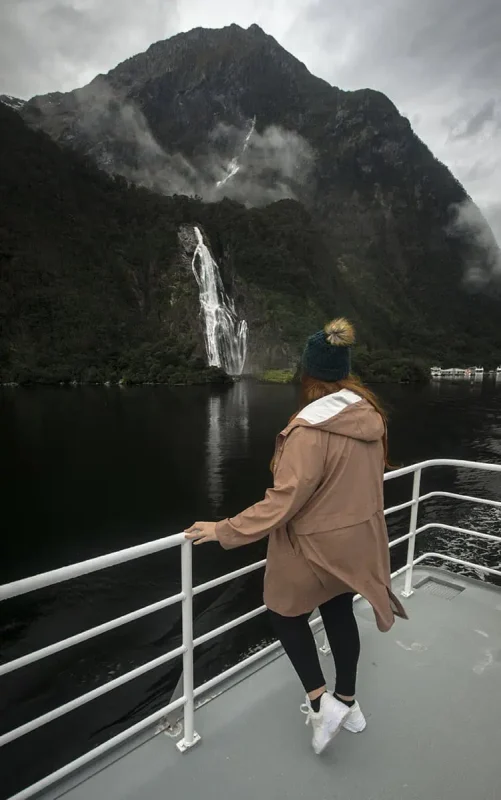 Bowen Falls In Milford Sound: The ULTIMATE Guide 2025 8 tasha amy in a brown coat and beanie stands on a boat deck, gazing at the majestic Bowen Falls in Milford Sound. The individual leans against the railing, captivated by the sight of the powerful waterfall cascading down the rugged, green mountainside. Mist from the falls drifts across the scene, blending with the clouds clinging to the mountain peaks, creating a serene and awe-inspiring backdrop.