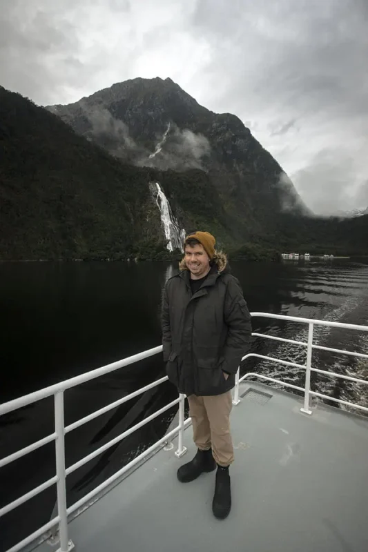 Bowen Falls In Milford Sound: The ULTIMATE Guide 2025 4 A person in a dark green parka with a fur-lined hood and a mustard yellow beanie stands on a boat deck in Milford Sound. The individual smiles as they pose against the railing, with the majestic Bowen Falls in the background, cascading down a steep, forested mountain. The overcast sky and the dark, reflective water enhance the serene and dramatic atmosphere of the scene.