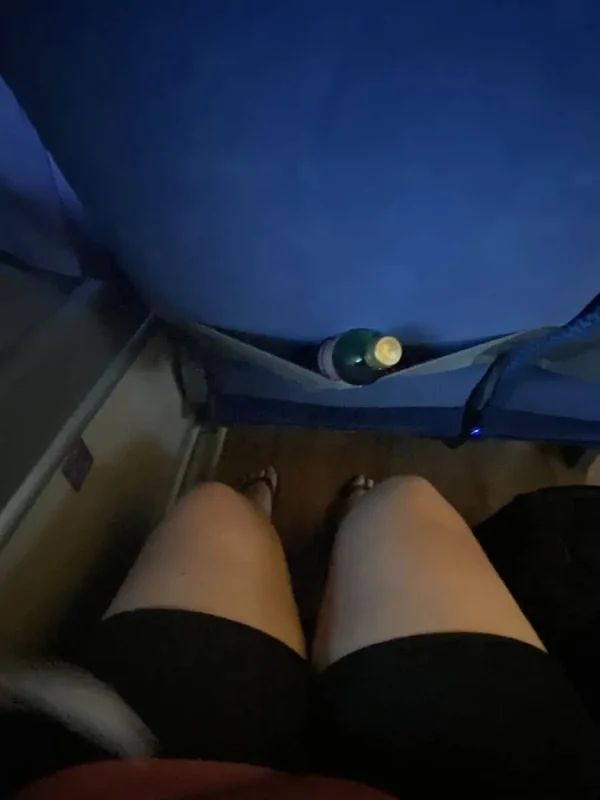 Cancun To Caye Caulker: The 4 Easiest Ways To Travel 7 A passenger's view inside a bus traveling from Cancun to Caye Caulker. The image shows a pair of knees in shorts, a bottle of water in the seat pocket, and a headrest with a paper cover labeled 'ADO'. The bus interior includes blue seats and ample legroom.