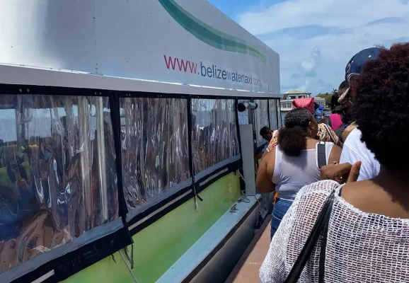 Passengers waiting in line to board the Belize Water Taxi ferry. The side of the ferry displays the website "www.belizewatertaxi.com" in bold letters. This vessel offers direct service from Chetumal to Caye Caulker, promising a scenic journey across the Caribbean Sea