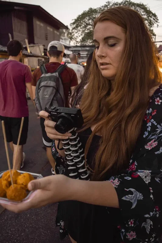 Chiang Mai Sunday Night Market: BEST Tips & Guide (2025) 5 Tasha Amy at the Sunday night market in Chiang Mai focused on her camera while holding a plate of Thai fried balls, with market-goers in the background.