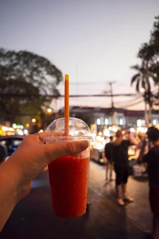 Chiang Mai Sunday Night Market: BEST Tips & Guide (2025) 4 Tasha holding a refreshing a strawberry smoothie with a bustling Sunday market Chiang Mai scene