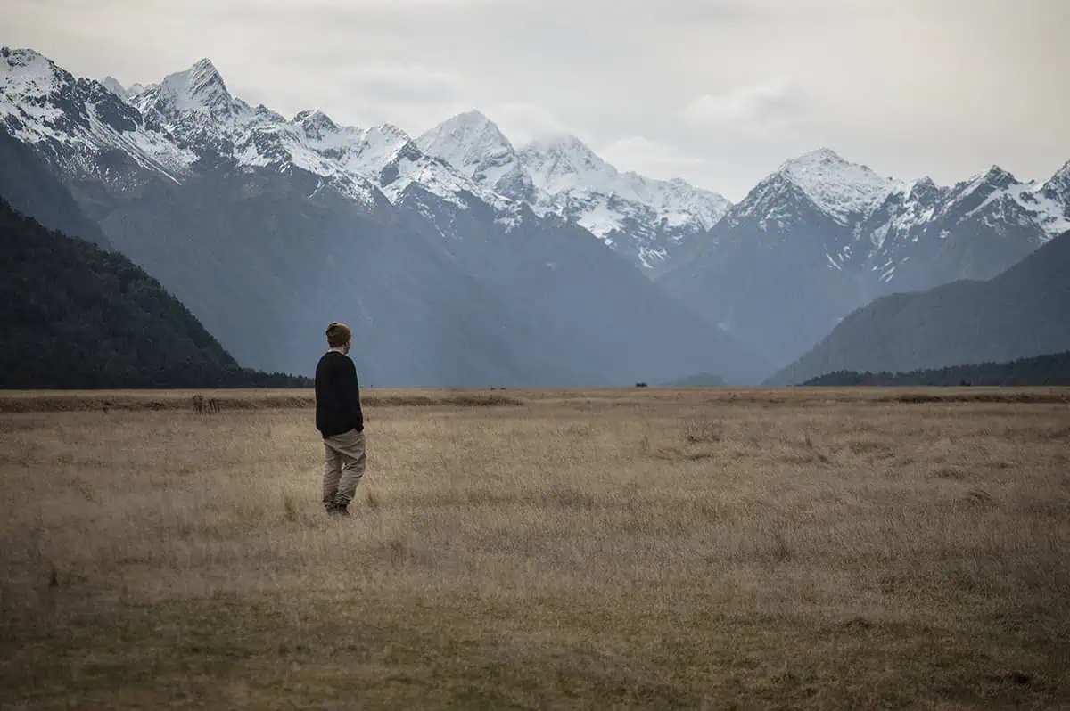 Eglinton Valley: ULTIMATE Guide For Visiting (2025) 9 A man gazes into the distance while standing in a wide expanse of dry grass, with towering snow-capped mountains creating a breathtaking backdrop. The tranquil setting of Eglinton Valley emphasizes the natural beauty of the area.