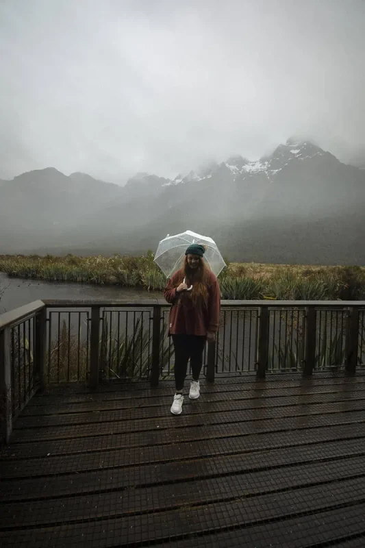 Eglinton Valley: ULTIMATE Guide For Visiting (2025) 16 tasha amy stands on a wooden platform holding a clear umbrella, with misty mountains and a river in the background. The overcast weather adds a mystical charm to the scenic landscape of Eglinton Valley.