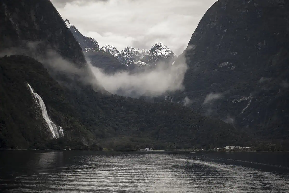 Eglinton Valley: ULTIMATE Guide For Visiting (2025) 23 A stunning view of a waterfall cascading down a lush green mountain into a calm lake, with mist and snow-capped peaks in the background. The majestic scenery highlights the natural splendor of milford sound
