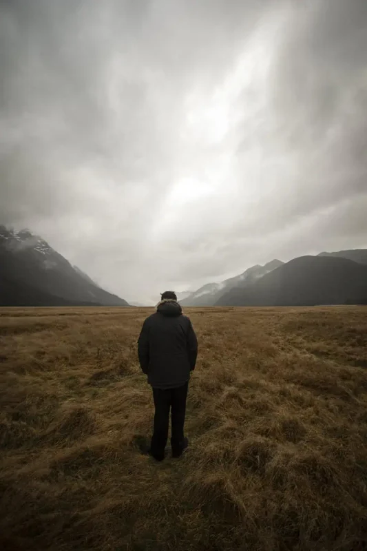 Eglinton Valley: ULTIMATE Guide For Visiting (2025) 14 A person in a dark coat stands facing away, looking towards the expansive plains and misty mountains in the distance. The overcast sky and muted tones create a reflective and tranquil mood in Eglinton Valley.